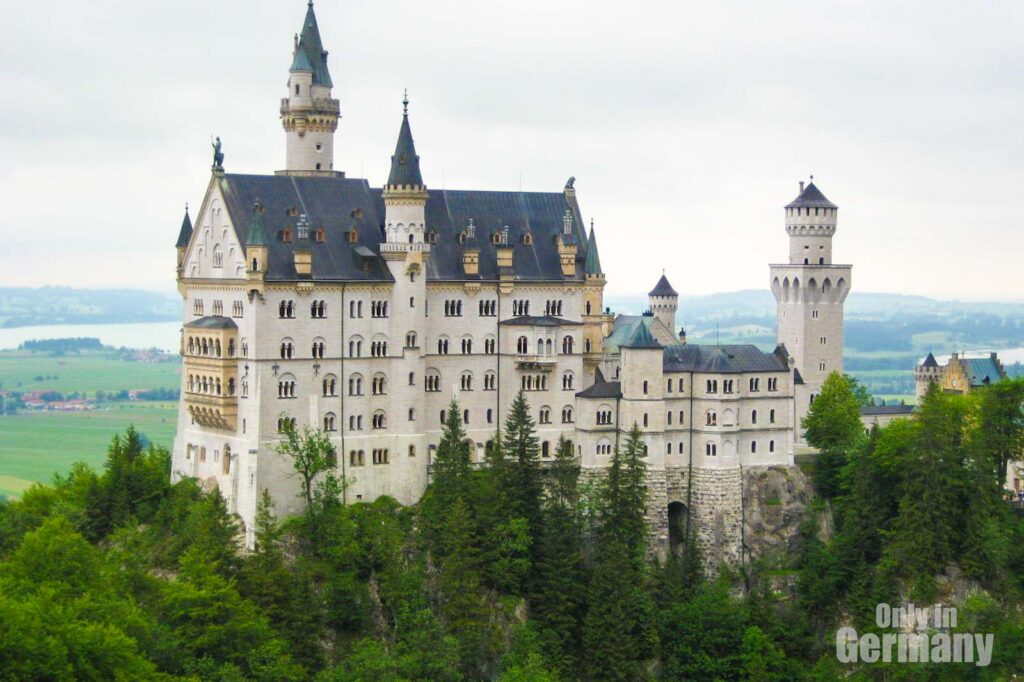 Neuschwanstein Castle in Germany seen from far away, surrounded by green trees.